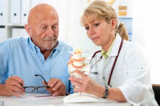 Doctor explains spinal anatomy to elderly man using a model.