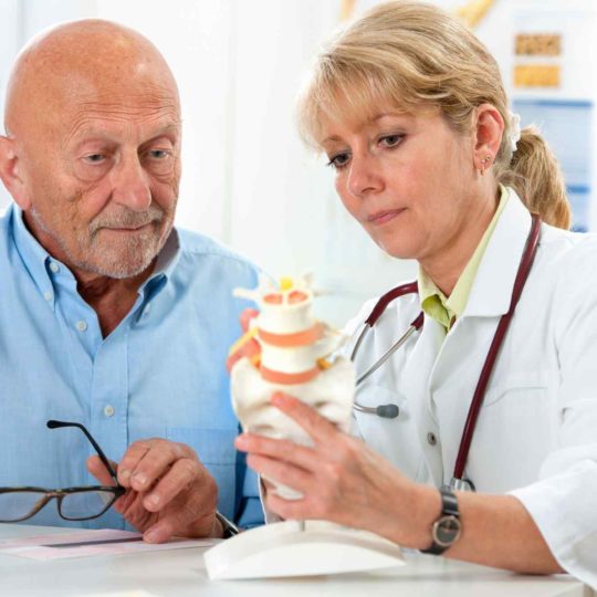 Doctor explains spinal anatomy to elderly man using a model.