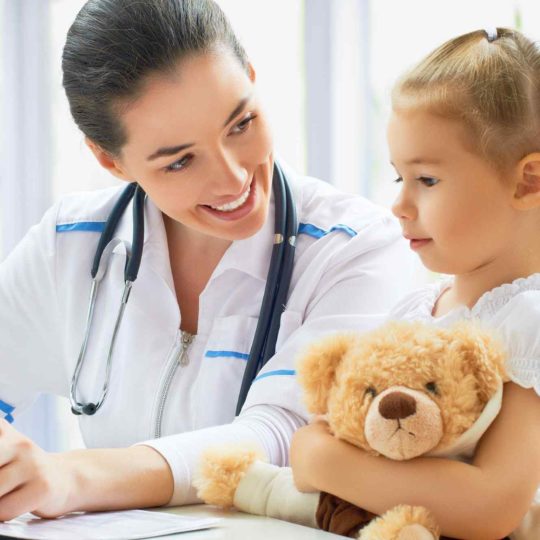 A female doctor smiling at a young girl holding a teddy bear.
