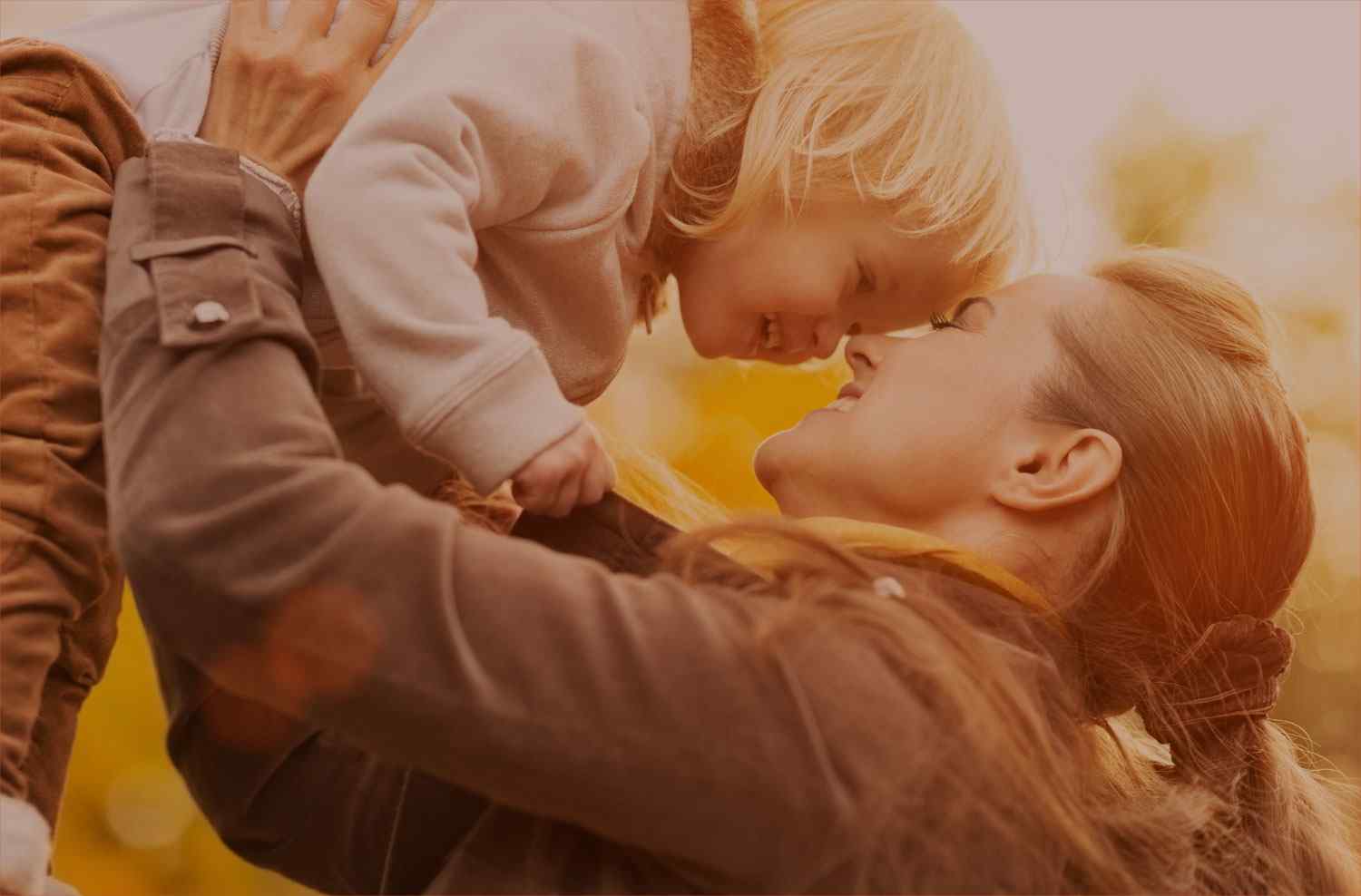 A mother joyfully lifts her baby in a warm, sunlit moment.